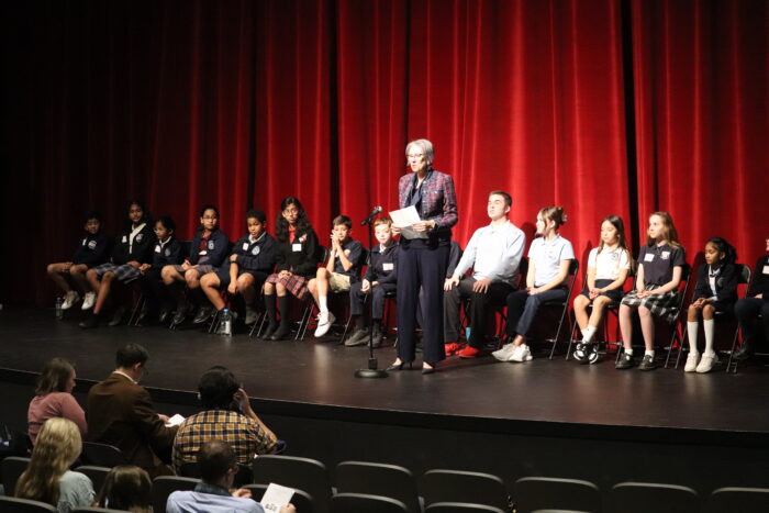 Students on chairs on a stage with Leanne Fawcett in front announcing the Spelling Bee